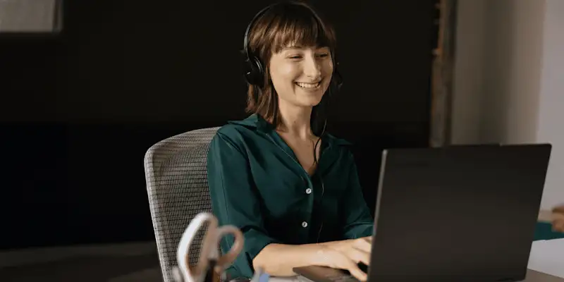 Smiling person wearing headphones and typing on a laptop computer at a desk in an office.