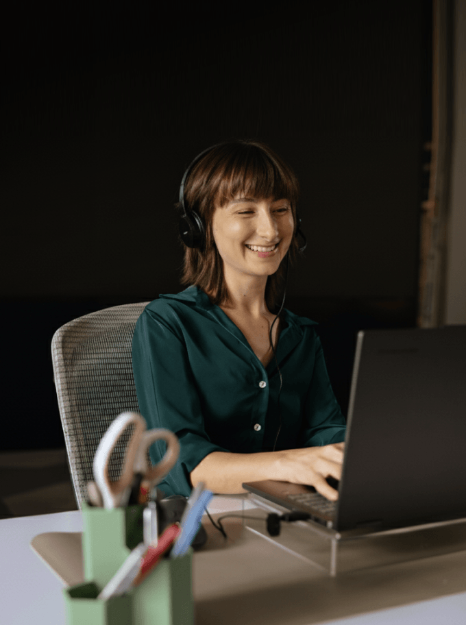 A smiling person wearing headphones works on a laptop at a desk with office supplies.