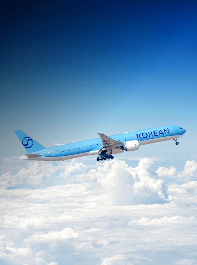 Korean Air Boeing 777 airplane ascending above clouds with a clear blue sky.