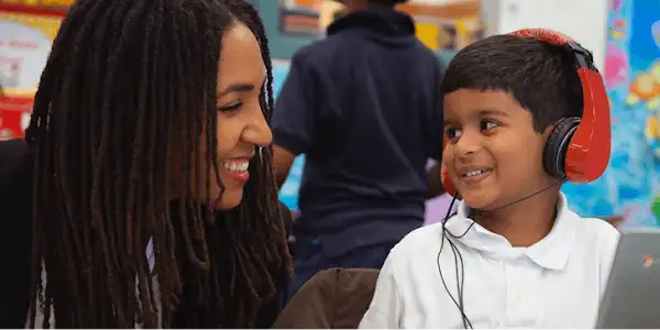 A teacher smiles while a young student wearing headphones looks at a laptop in a classroom setting.