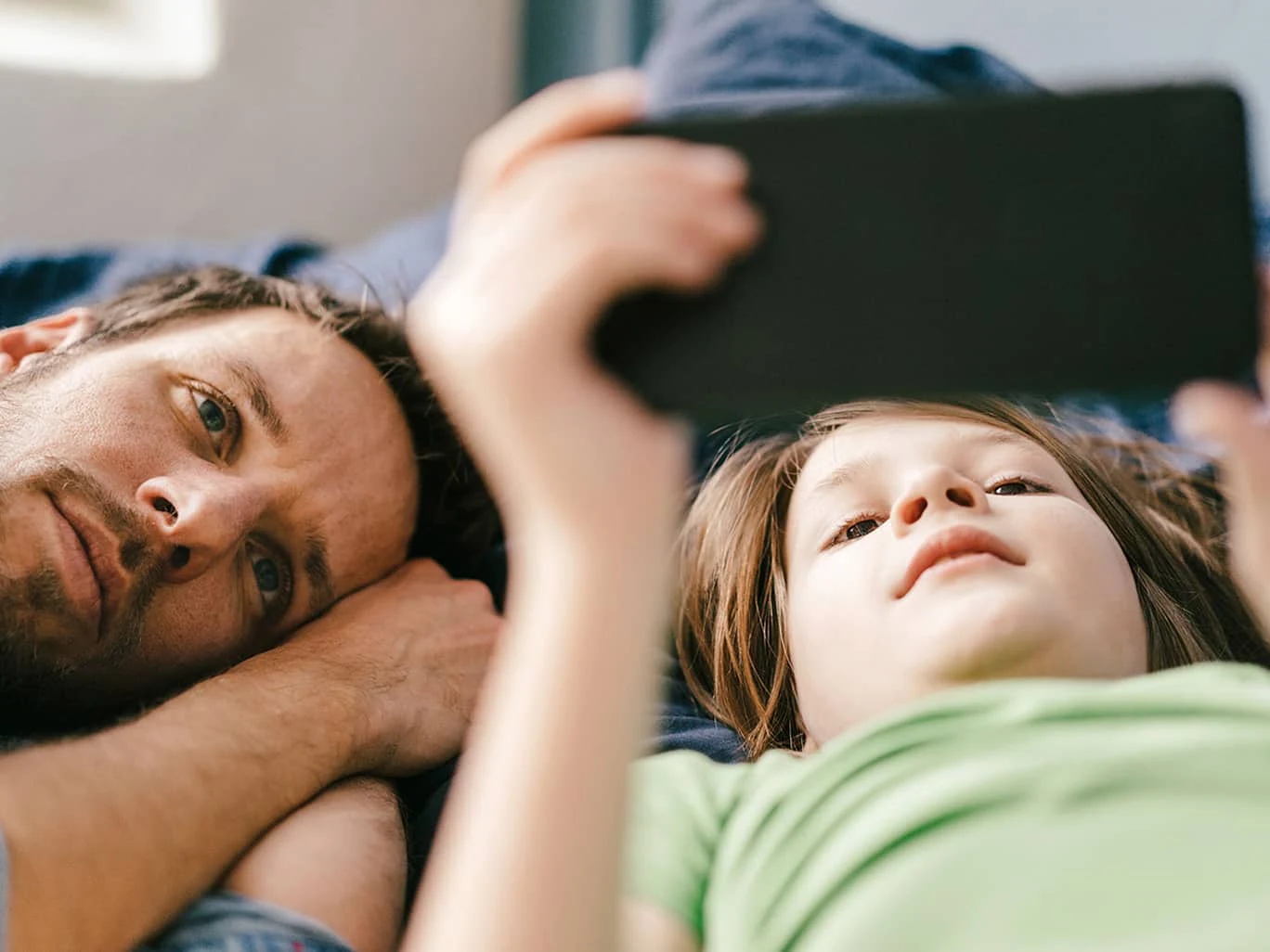 A child and an adult are lying down on the bed. They both watch something on a device.