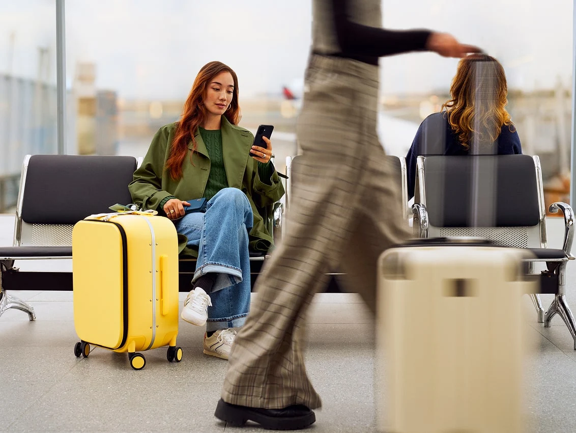 An individual sitting in an airport with a yellow suitcase, with other people walking through the airport with luggage.