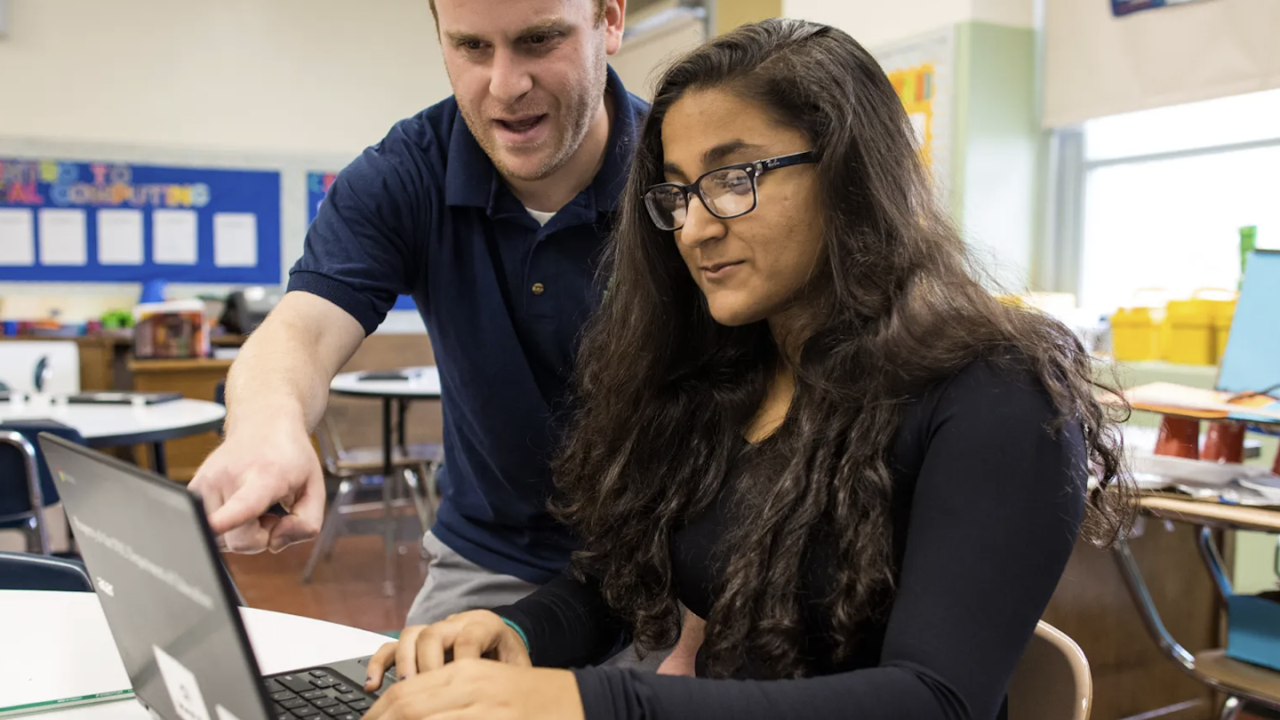 A male teacher wearing a navy blue polo shirt leans over to point at a laptop screen, guiding a female student who is seated and typing. The student has long dark wavy hair, wears glasses, and is dressed in a black long-sleeved shirt. They are in a brightly lit classroom setting with a bulletin board, circular tables, and chairs visible in the softly blurred background.