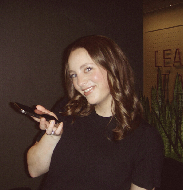 A smiling woman with wavy brown hair holds a smartphone in one hand while standing indoors. She wears a black top, and green plants and a partial wall sign are visible in the background.