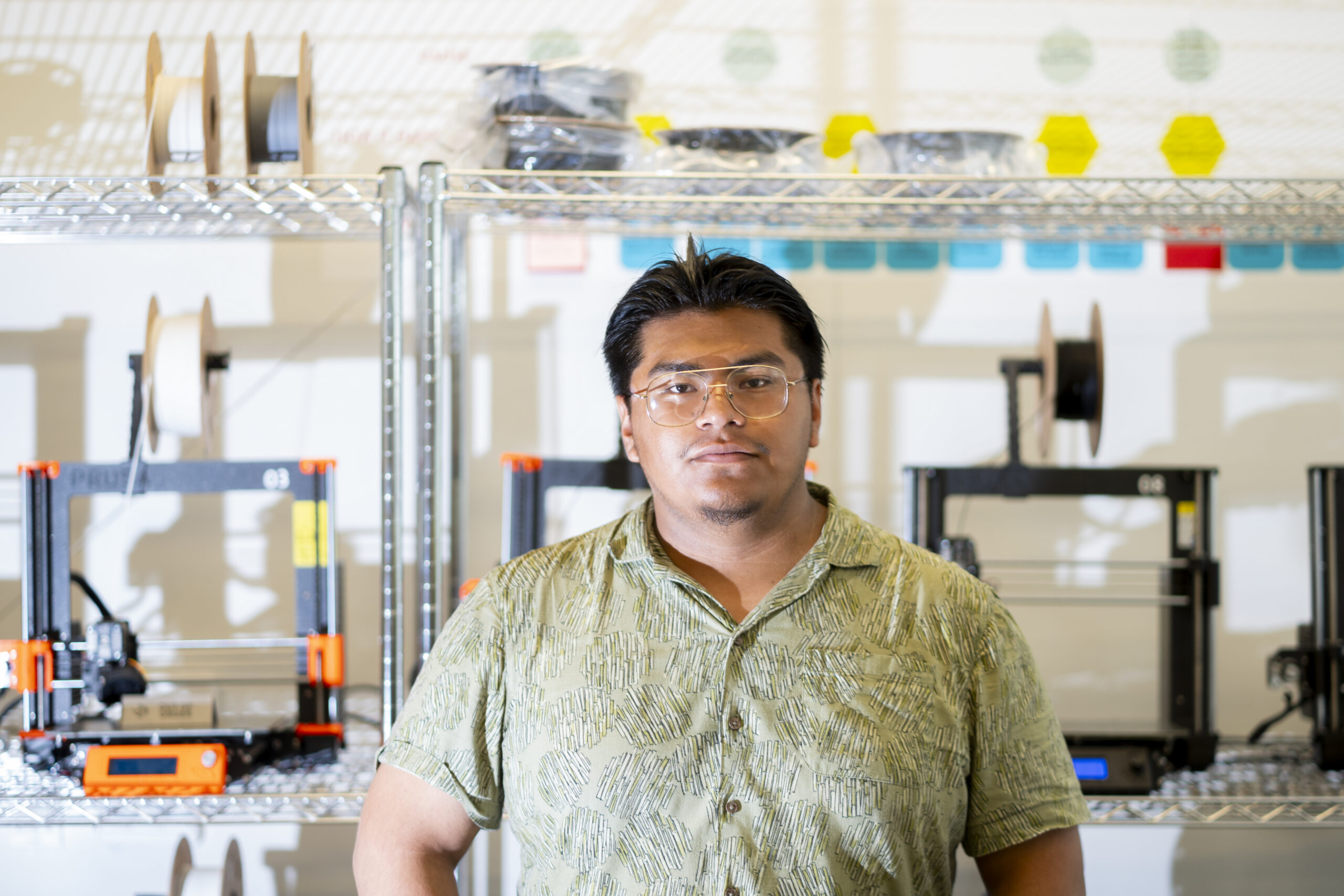 Portrait of Israel Sanchz standing in front of wire shelves of engineering equipment wearing a light green short-sleeve button-down shirt and wire-rimmed glasses
