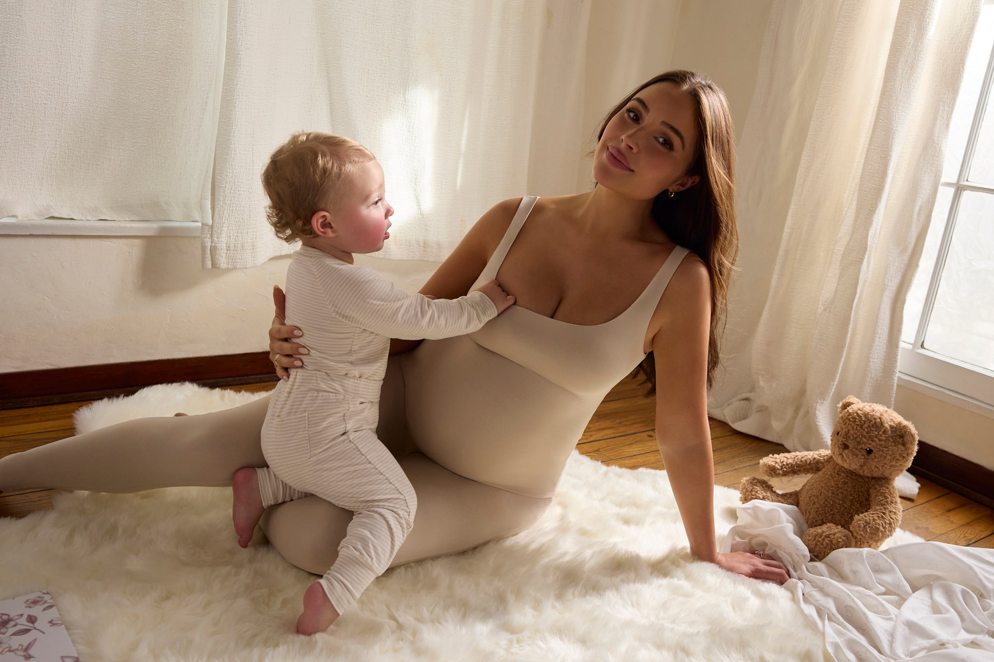 Olivia Culpo sits on a white rug with a young child on her lap, wearing a matching beige outfit.