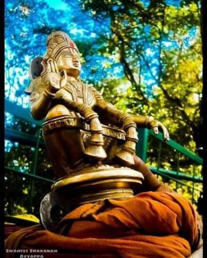 Golden Ayyappa statue draped in orange cloth surrounded by green trees. — God Wallpaper