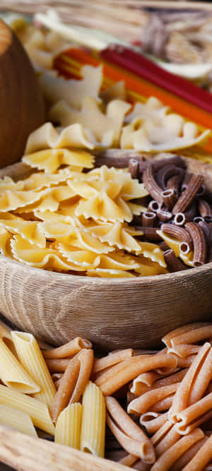 A wooden bowl filled with assorted uncooked pasta shapes. — OnePlus 11 Wallpaper