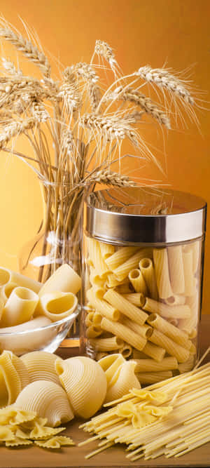 Glass jar and bowl filled with various types of uncooked pasta beside wheat stalks on wooden surface. — OnePlus 11 Wallpaper