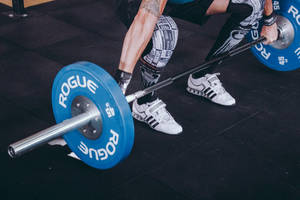 Weightlifter performing squat with blue Rogue barbell on gym floor. — Barbell Wallpaper