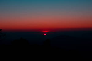 Um sol vermelho se pondo atrás de silhuetas de montanhas em um céu gradiente. — Relaxe Papéis de Parede