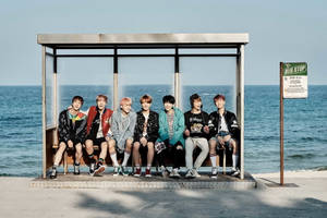 Seven young men in trendy outfits sitting at a seaside bus stop. — Bts Wallpaper