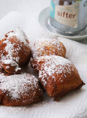 Gyldne beignets dækket med flormelis på et hvidt håndklæde med en sigte i baggrunden. — Design Wallpapers