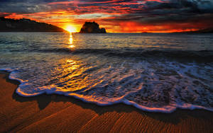 Tramonto sull'oceano con onde che lambiscono la sabbia e isola rocciosa lontana — HD Sfondi