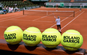 Quattro palline da tennis Roland Garros su un bordo di legno con un campo in terra battuta e un giocatore in movimento sullo sfondo. — Francese aperto Sfondi
