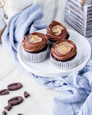 Tres cupcakes de chocolate con pan de oro sobre un plato blanco con paño azul y trozos de chocolate. — Cupcake Fondos de pantalla
