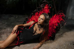 Uma mulher com asas de penas vermelhas e pretas deitada em solo arenoso numa floresta escura. — Moda Papéis de Parede