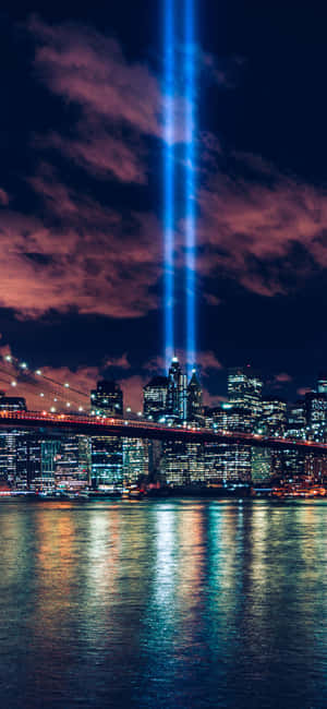 Two blue tribute lights shine over a city skyline at night reflected in water. — iPhone 13 Mini Wallpaper