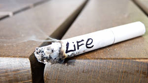 A burning cigarette labeled 'Life' rests on a wooden surface with smoke rising. — Death Wallpaper