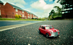 Um Mini Cooper de brinquedo vermelho em uma rua com casas e árvores ao fundo. — Carro Papéis de Parede