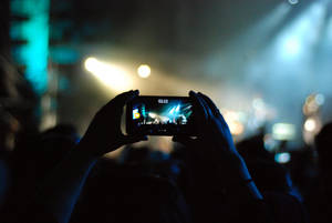 Hender holder telefon og spiller inn en livekonsert under fargerike scenelys. — Mobile Bakgrunner