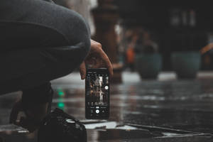 Person på huk for å ta et bilde av en regnfull bygate med telefon — Mobile Bakgrunner