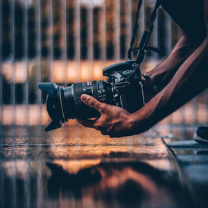 Handen houden een DSLR-camera boven nat trottoir dat gouden stadslichten reflecteert in de schemering. — Camera Achtergronden