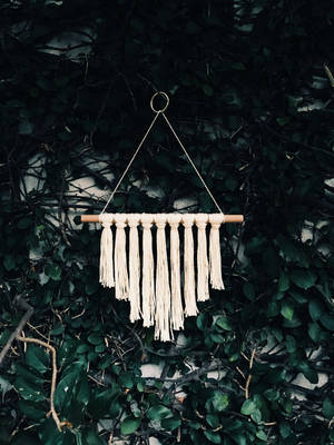 Cream macrame wall hanging with wooden dowel and tassels against green foliage. — iPhone 7 Plus Wallpaper