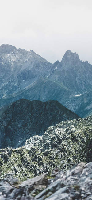 Ruggede bjergtoppe rejser sig gennem tåge med klippefyldt forgrund. — OnePlus Wallpapers
