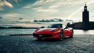 Ferrari vermelha estacionada em paralelepípedos perto da água com torre histórica ao fundo ao pôr do sol. — Carro Papéis de Parede