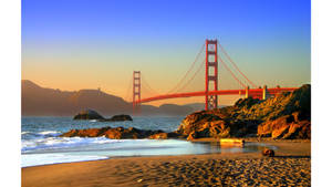 Golden Gate Bridge at sunset over a sandy beach with waves and rocks. — Travel Wallpaper