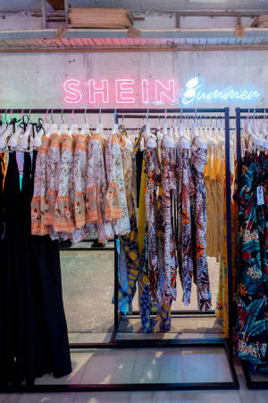 Vestidos de verano coloridos exhibidos bajo un letrero de neón de SHEIN en una tienda de ropa. — She Fondos de pantalla
