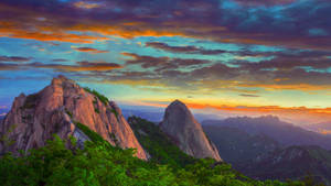 Cordilheira dramática banhada em cores de pôr do sol com árvores verdes vibrantes em primeiro plano. — Tranquilo Papéis de Parede