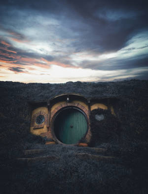 Maison de hobbit avec porte ronde couleur sarcelle dans une colline herbeuse au crépuscule.