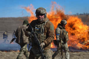 Soldiers in combat gear advancing through a field with a large fire behind them. — War Wallpaper