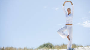 Woman in white yoga outfit balancing in tree pose on a beach dune under blue sky. — Yoga Wallpaper