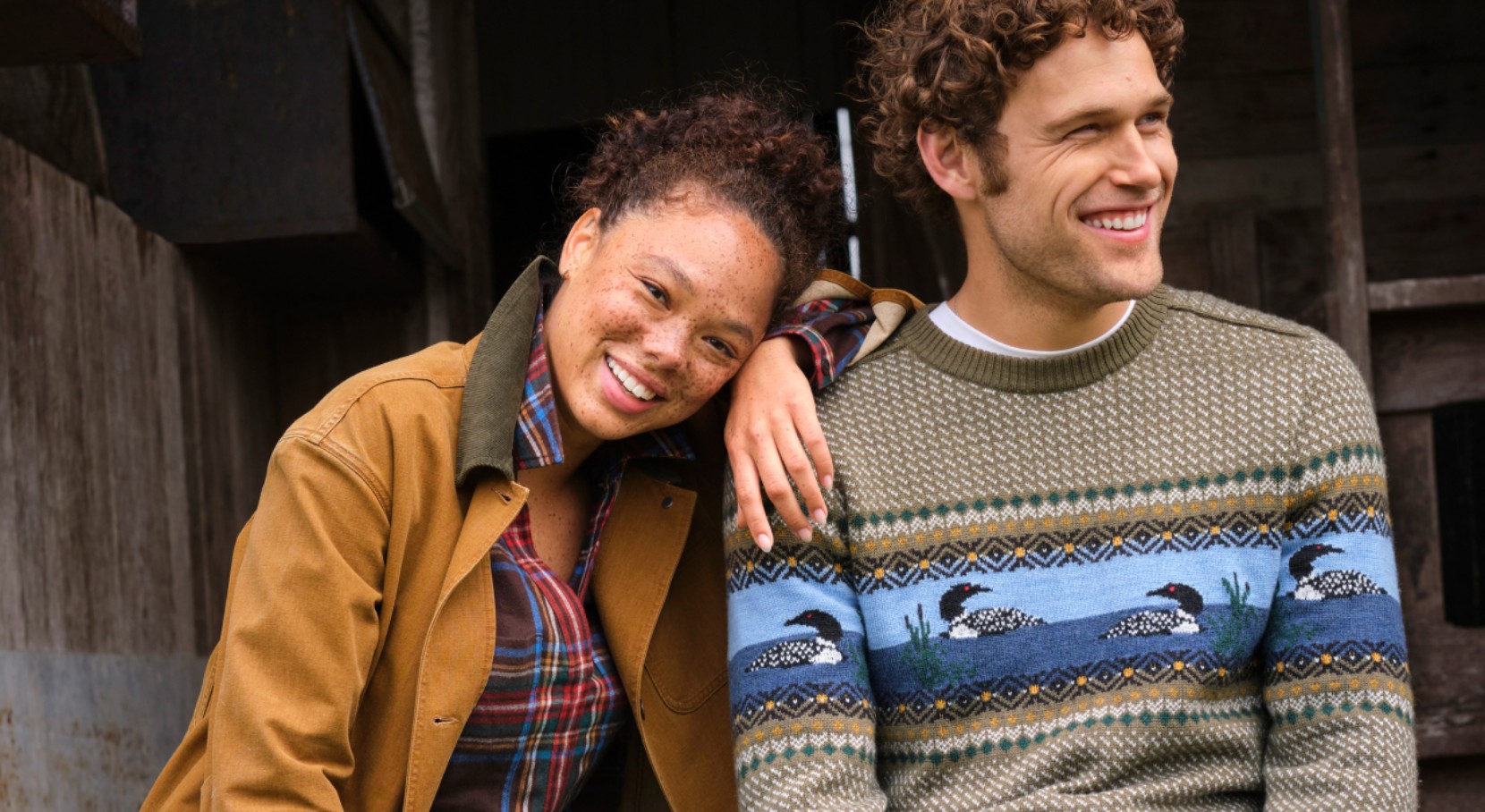 Two people in fall layers sit outside a rustic building; one wears a loon-pattern sweater and the other a plaid shirt and jacket.