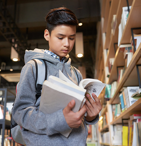 Image shows a boy standing in a library reading a book.