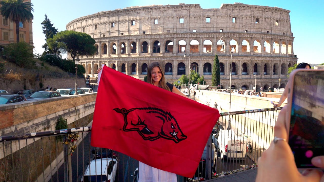 Student displaying a red Razorback flag in front of the historic Colosseum in Rome.